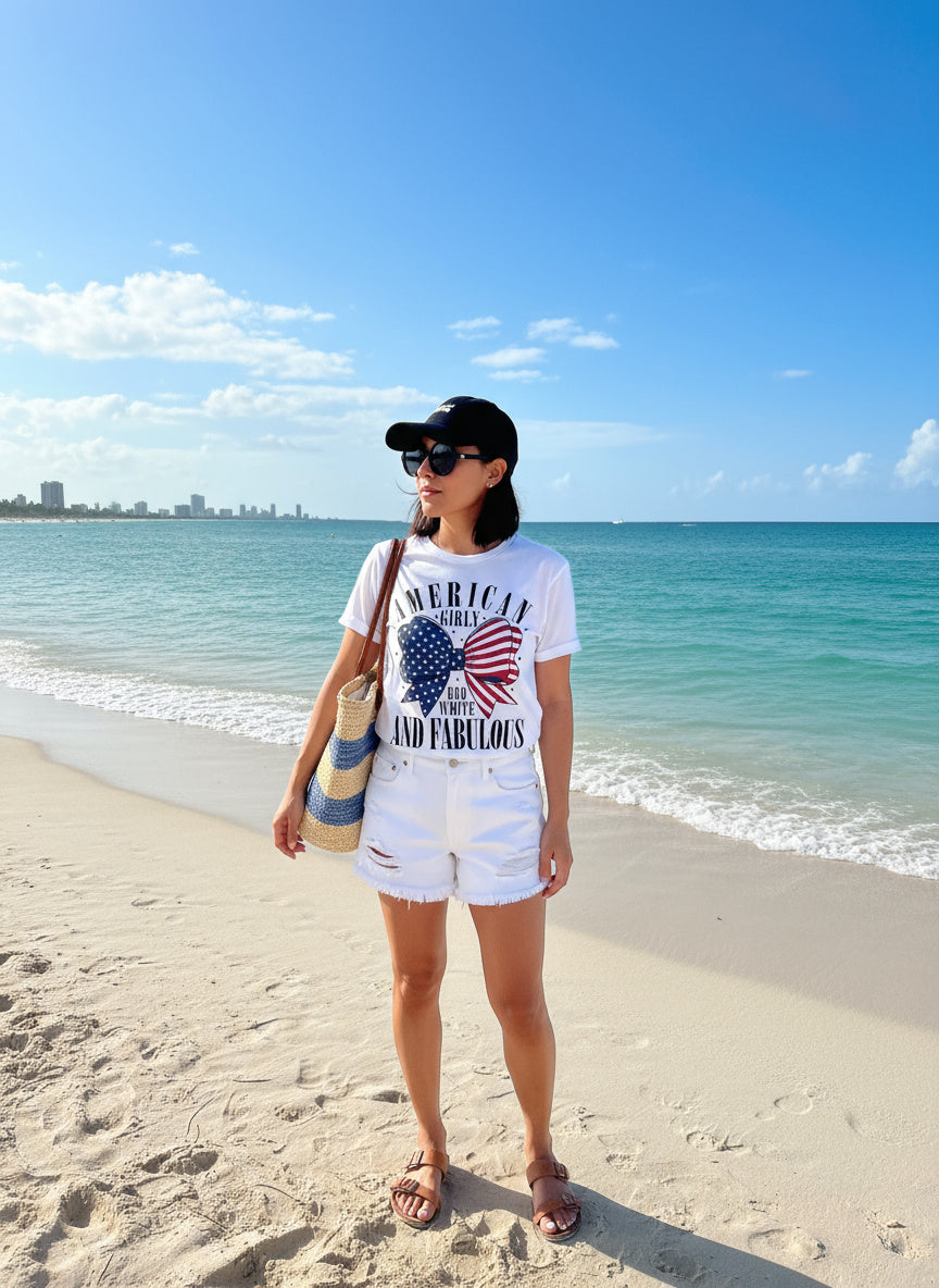 Woman standing on a beach wearing a white t-shirt with text and graphics, shorts, and a cap.